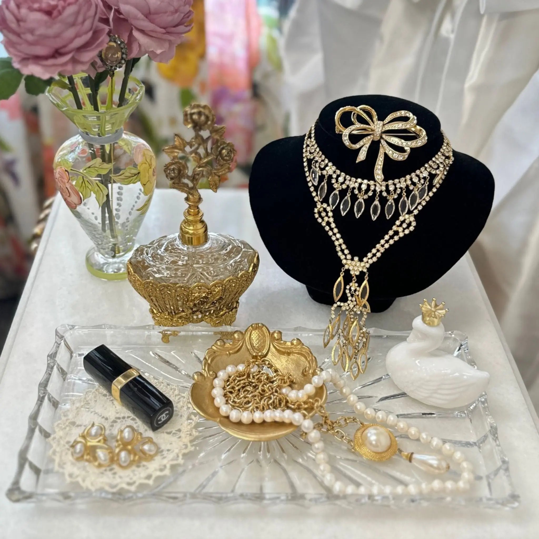 Romantic bridal jewelry display with crystal bow necklace, layered pearls, and gold vintage accessories styled with roses and antique vanity décor.