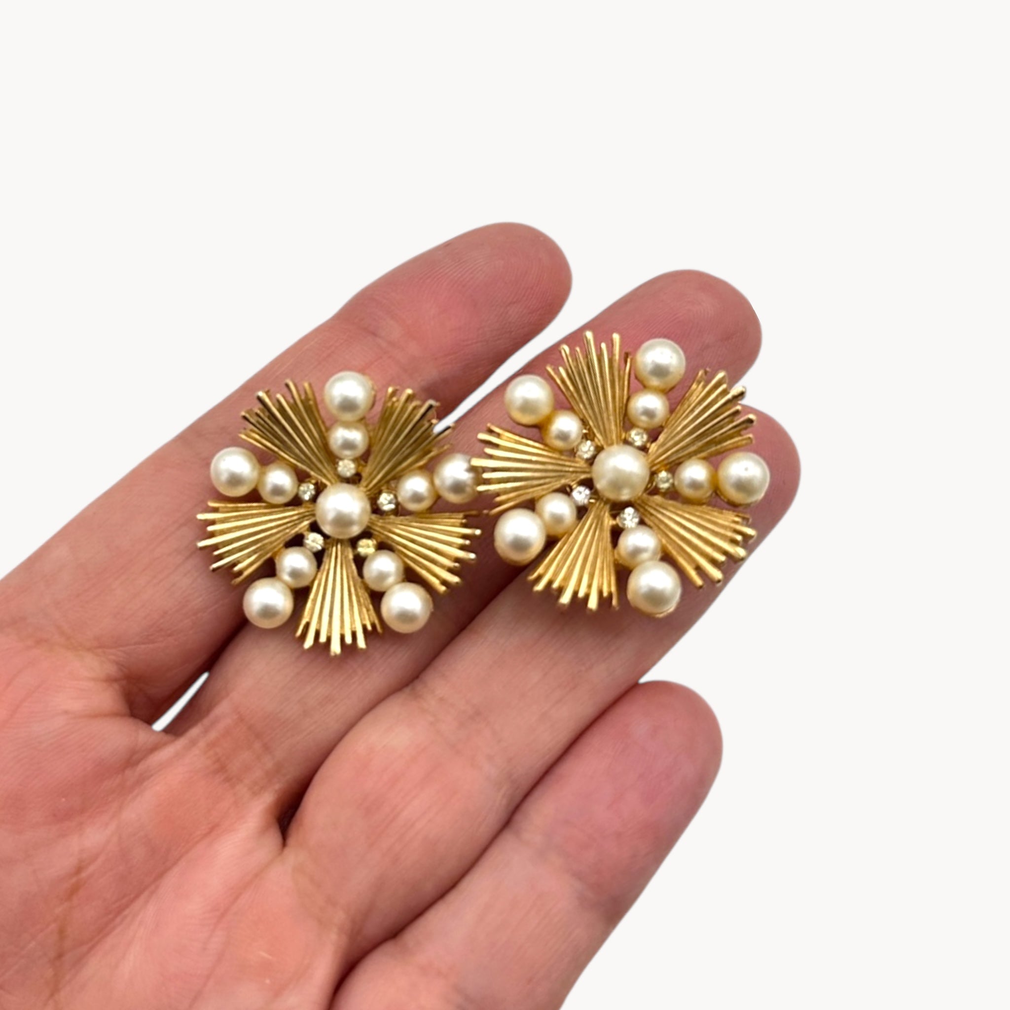 Scale size view of Trifari gold starburst clip-on earrings with faux pearls and rhinestones, 1960s jewelry on white background.