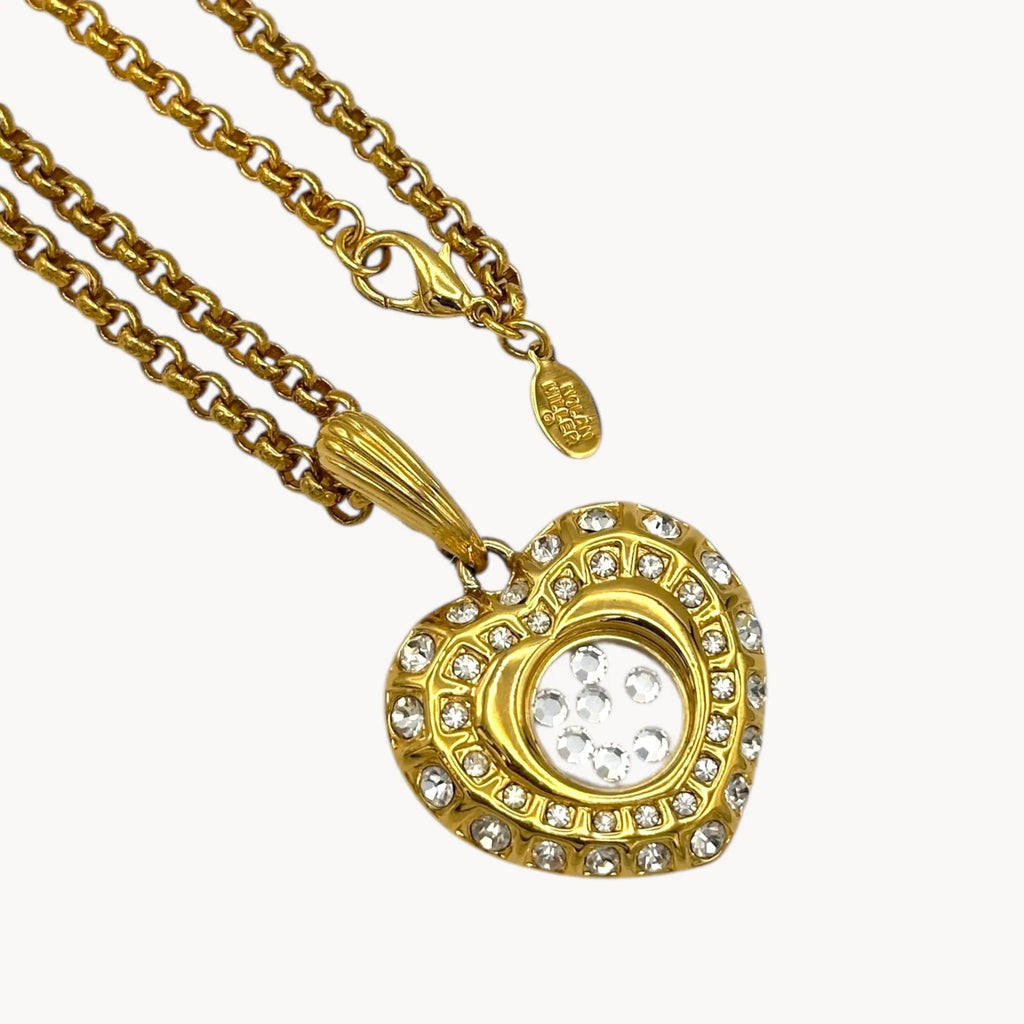 Close up of pendant and hang tag of Nolan Miller gold-tone heart pendant necklace with clear rhinestones on long chain on white background.