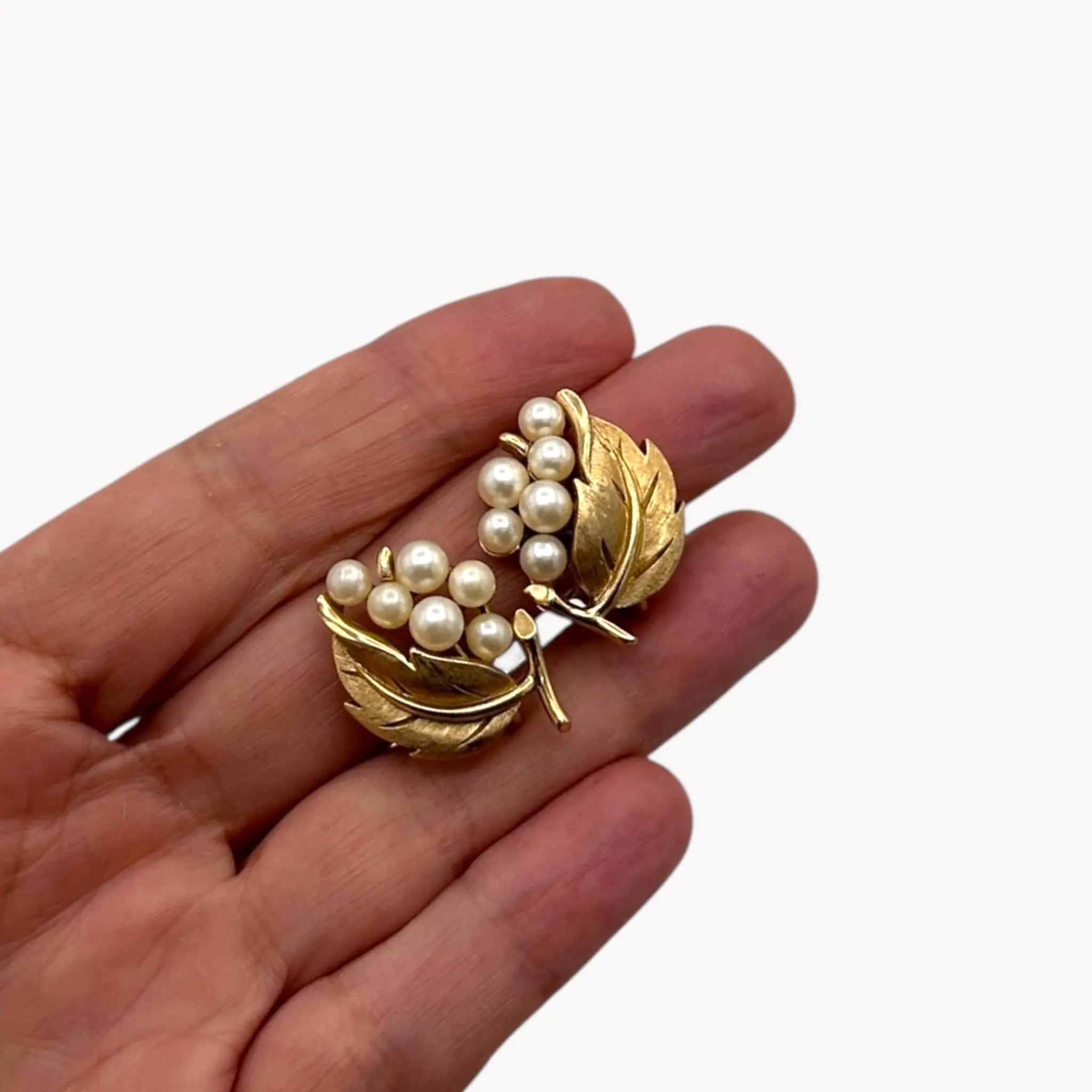 On hand Vintage Trifari gold leaf clip-on earrings with faux pearl clusters, mid-century jewelry on white back background.