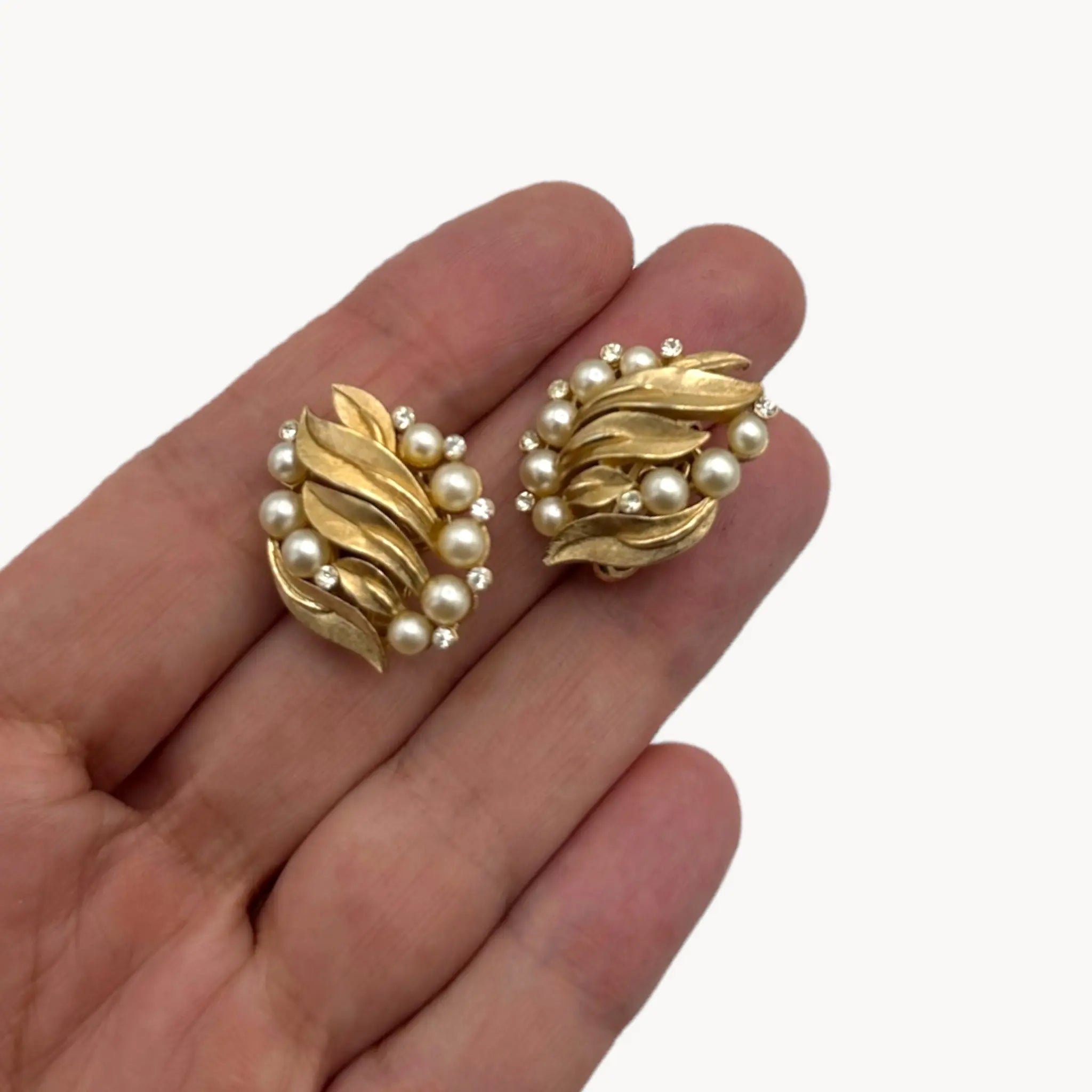 Scale size view on hand of Vintage Trifari gold floral clip-on earrings with faux pearls and rhinestones, 1960s jewelry on white background.