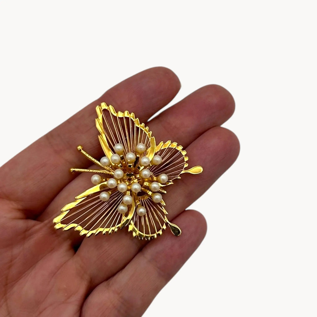 Scale size view on hand of Vintage Monet butterfly brooch with gold openwork wings and faux pearl accents on white background.
