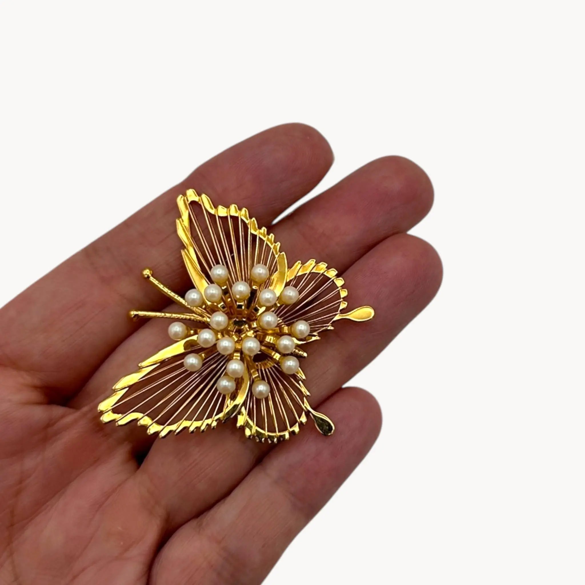 Scale size view on hand of Vintage Monet butterfly brooch with gold openwork wings and faux pearl accents on white background.