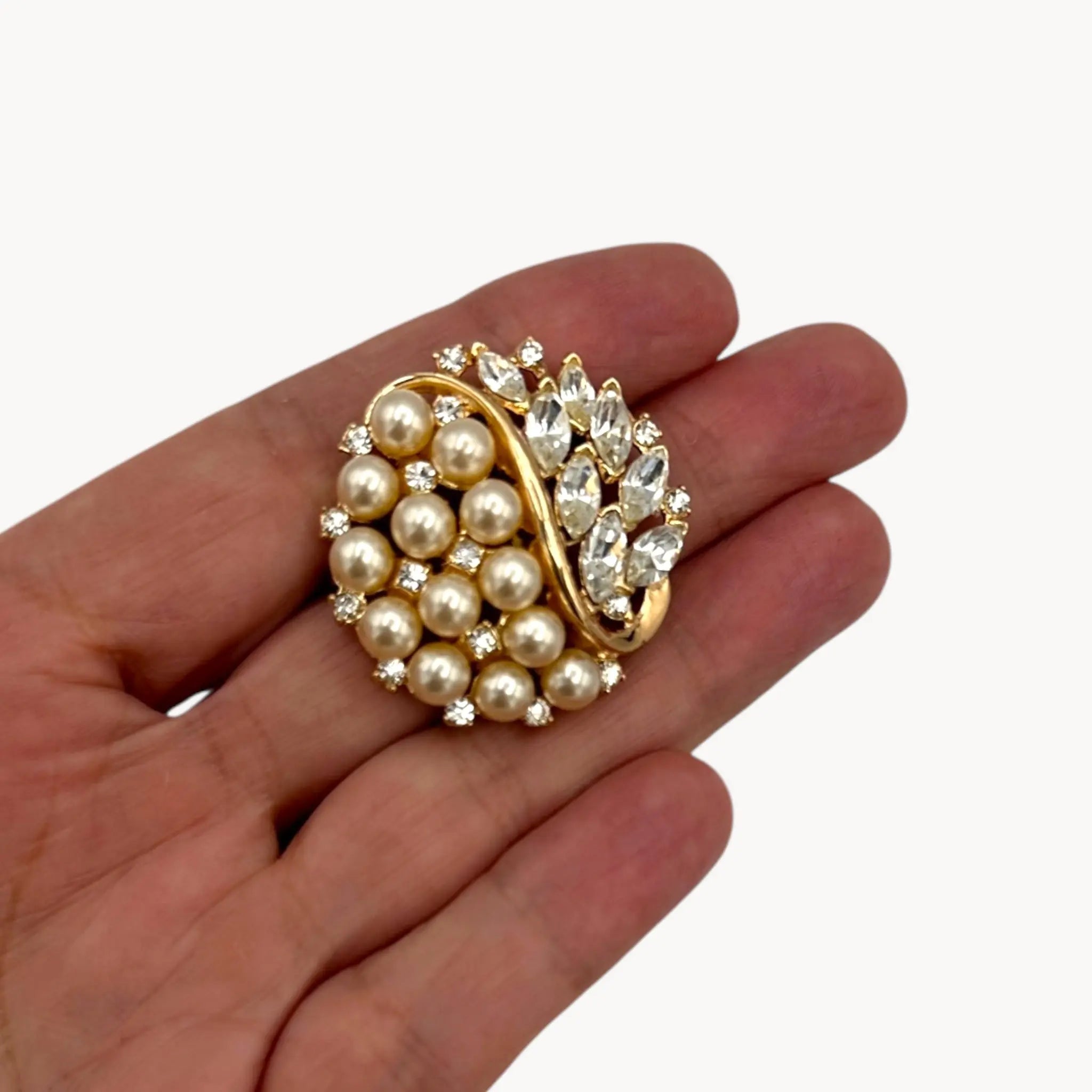 Brooch on hand of the Vintage Trifari brooch with faux pearls, rhinestones, and gold-tone design shown on white background.